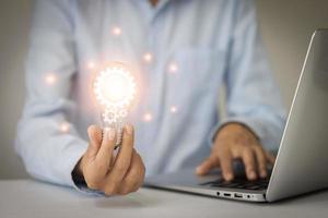 The hand of a man with a light bulb And there is a gear icon in it And he is using a notebook computer. photo