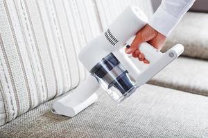 A person uses a white cordless vacuum cleaner to clean the sofa in the living room. photo