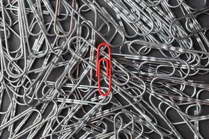 A red paper clip stands out against a textured background of silver paper clips