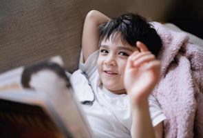 Happy School kid lying on sofa showing hand and looking up with  smiling face, Child boy reading story relaxing at home on weekend, Positive children concept photo