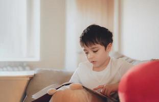 niño de la escuela feliz sentado en el sofá leyendo un libro con la luz de la mañana brillando desde la ventana. niño niño leyendo historia relajándose en casa el fin de semana, concepto de niños positivos foto