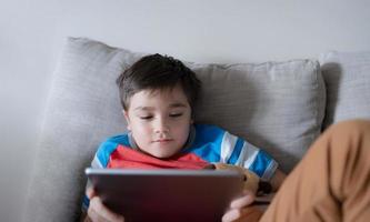 School kid using tablet readying story from internet, Happy boy sitting on sofa doing studying online learning  at home, Child holding digital pad using wifi playing game or chatting with friends. photo