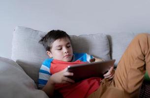 School kid using tablet readying story from internet, Happy boy sitting on sofa doing studying online learning  at home, Child holding digital pad using wifi playing game or chatting with friends. photo