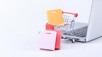 Online shopping. Mini shop cart trolley with colorful paper bags over a laptop computer on white table background, buying at home concept, close up photo