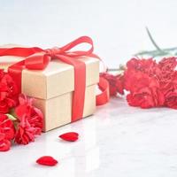 Mother's Day gift background, red carnation flower bouquet with wrapped kraft gift tied with ribbon isolated on marble white background, close up. photo