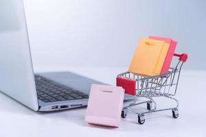 Online shopping. Mini shop cart trolley with colorful paper bags over a laptop computer on white table background, buying at home concept, close up photo