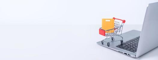 Online shopping. Mini shop cart trolley with colorful paper bags over a laptop computer on white table background, buying at home concept, close up photo