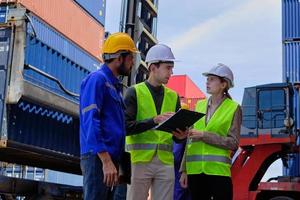 Three professional Caucasian workers team in safety uniforms and hardhats work at a logistics crane with many stacks of containers, loading control shipping goods for cargo transportation industry. photo