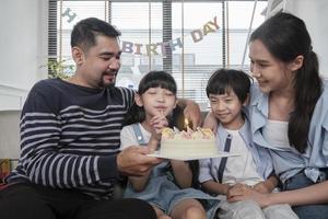 Happy Asian Thai family, young daughter is surprised with birthday cake, blows candle, pray and cheerful celebrates party with parents together in living room, wellbeing domestic home event lifestyle. photo