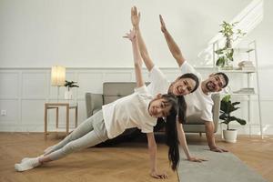 Asian Thai parents and daughter fitness training exercise and practice yoga on living room floor, lovely rowing together for health and wellness, and happy domestic home lifestyle on family weekend. photo