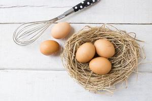 Fresh organic eggs on nest with whisk on white wooden background. photo