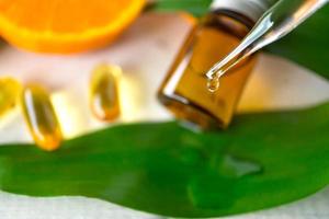 Vitamin C serum dropping from droper with a bottle, capsule, green leaf and orange background. Selective focus. photo