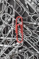 A red paper clip stands out against a textured background of silver paper clips