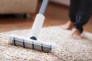 piernas humanas y un cepillo turbo blanco de una aspiradora inalámbrica limpia la alfombra de la casa foto