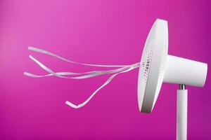 The electric fan is white with pink ribbons fluttering in the wind on a pink background. photo