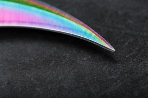 The curved sharp blade of the Kerambit Dagger is a gradient rainbow color on a dark background. photo