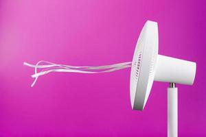 The electric fan is white with pink ribbons fluttering in the wind on a pink background. photo