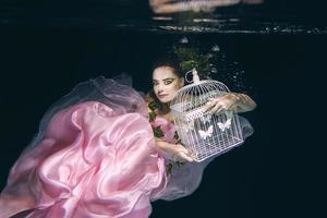 young beautiful woman in long  pink dress with cage in her hands underwater photo