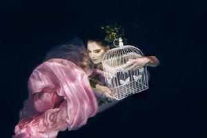 young beautiful woman in long  pink dress with cage in her hands underwater photo