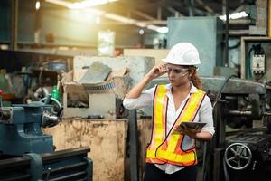 Female industrial engineer wearing a white helmet while standing in a heavy industrial factory behind she looking of working at industrial machinery and check security system setup in factory. photo
