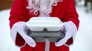 Lebensmittel-Lieferservice-Container in den Händen des Weihnachtsmanns im Freien im Schnee. Weihnachtsaktion. fertige heiße Bestellung, Einweg-Plastikbox. Silvester Catering. Platz kopieren, verspotten