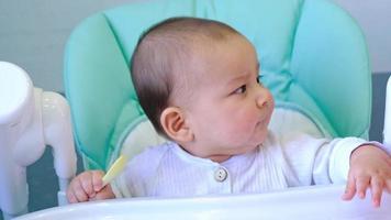 A hungry baby is gnawing on a plastic spoon at the table on a high chair. Teething, whims, itchy gums, introduction of complementary foods video