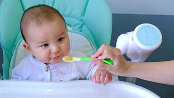 Mom feeds the baby with a spoon of vegetable puree at the children's feeding table. Baby's appetite, healthy nutrition, introduction of complementary foods. Copyspace, mock up video