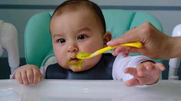 Mom feeds the baby with a spoon of vegetable puree at the children's feeding table. Baby's appetite, healthy nutrition, introduction of complementary foods. Copyspace, mock up video