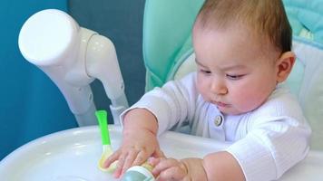 A hungry baby is gnawing on a plastic spoon at the table on a high chair. Teething, whims, itchy gums, introduction of complementary foods video