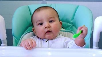 A hungry baby is gnawing on a plastic spoon at the table on a high chair. Teething, whims, itchy gums, introduction of complementary foods video