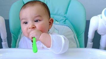 un bebé hambriento está mordiendo una cuchara de plástico en la mesa de una silla alta. dentición, caprichos, picor de encías, introducción de alimentos complementarios video