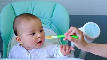 Mom feeds the baby with a spoon of vegetable puree at the children's feeding table. Baby's appetite, healthy nutrition, introduction of complementary foods. Copyspace, mock up video