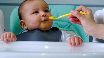 Mom feeds the baby with a spoon of vegetable puree at the children's feeding table. Baby's appetite, healthy nutrition, introduction of complementary foods. Copyspace, mock up video