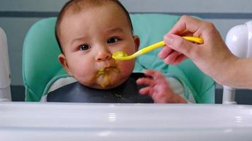 Mom feeds the baby with a spoon of vegetable puree at the children's feeding table. Baby's appetite, healthy nutrition, introduction of complementary foods. Copyspace, mock up video