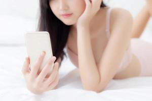 Closeup beautiful young asian woman sexy in underwear chatting on mobile phone at bedroom, happy girl in lingerie looking social media on smartphone lying on bed, communication and lifestyle concept. photo