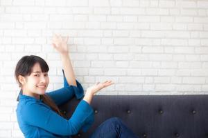 hermosa joven asiática sentada y sonriendo feliz y presentando algo en el sofá mostrando el futuro o planeando con un mensaje tuyo, chica alegre y relajada en el sofá en casa, concepto de estilo de vida. foto