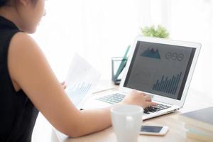 Young asian woman working and typing with laptop computer at office, businesswoman looking graph diagram analysis statistic data of finance, girl using notebook, profit growth, business concept. photo