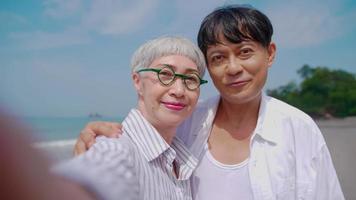 Asian retired couple relaxing by the sea in summer. A senior woman taking a selfie photo with her partner on the beach.