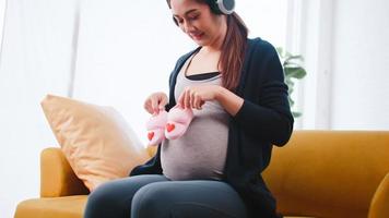 Happy Asian pregnant woman listening to music with headphones on sofa at home. photo