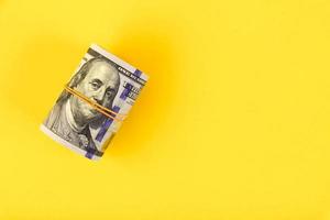 A roll of hundred-dollar American bills is tied with elastic band on a yellow background. Copy space photo