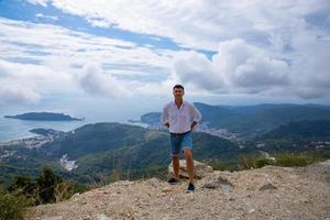 Caucasian man posing on the edge of a mountain peak with a gorgeous view of the Adriatic Sea and the city of Budva, Montenegro. photo