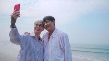 Asian retired couple relaxing by the sea in summer. A senior woman taking a selfie photo with her partner on the beach.