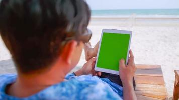 Asian man watching something on his tablet while relaxing on the beach. photo