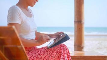 Asian woman drawing on a tablet while sitting by the sea. Freelancer working on the beach. photo