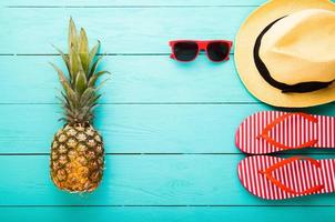 Flip flops, pineapple, hat and sunglasses. Summer accessories for modern woman on her vacation. Top view. Blue wooden background with copy space. photo
