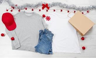 cierre la camiseta blanca con plantilla en blanco, sudadera con capucha con espacio para copiar. concepto de vacaciones de navidad. camiseta de bolsa de papel de maqueta de vista superior, gorra roja sobre fondo de madera blanca. feliz año nuevo decoraciones foto