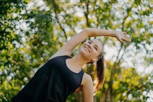 la mujer de fitness deportivo se calienta y los ejercicios de estiramiento se inclinan a un lado tiene una expresión facial contenta vestida con poses de ropa activa contra el fondo de la naturaleza durante el día de verano. deporte y motivacion foto