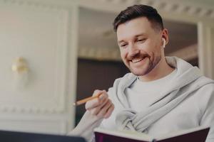 Smiling man freelancer in pullover and wireless earphones with pencil and book in hands having video call on laptop photo