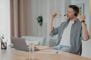 Happy successful man clenches fists celebrates triumph works online wears headset poses at desktop with laptop computer dressed casually. Overjoyed male student listens educational online lecture photo