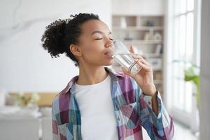 una joven afroamericana disfruta bebiendo agua pura filtrada de vidrio en casa. estilo de vida saludable foto